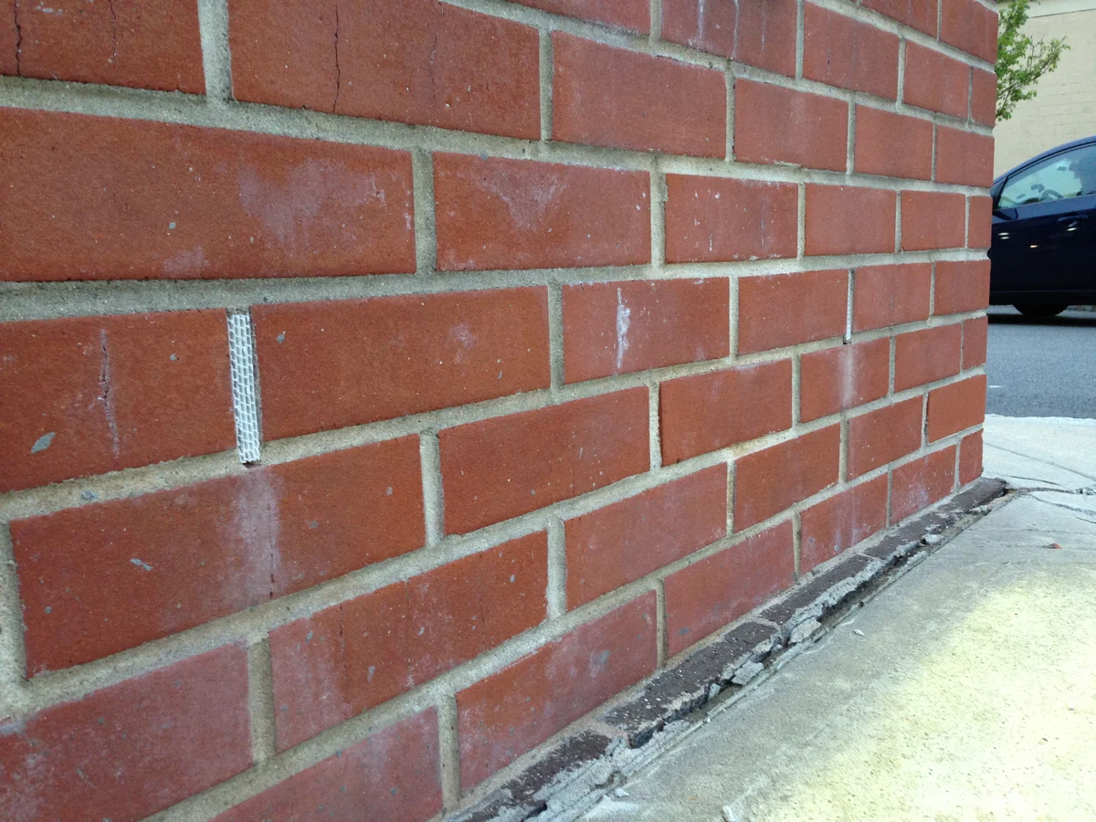 Red brick wall with a narrow vertical weep hole fitted with a white plastic grid insert near the base