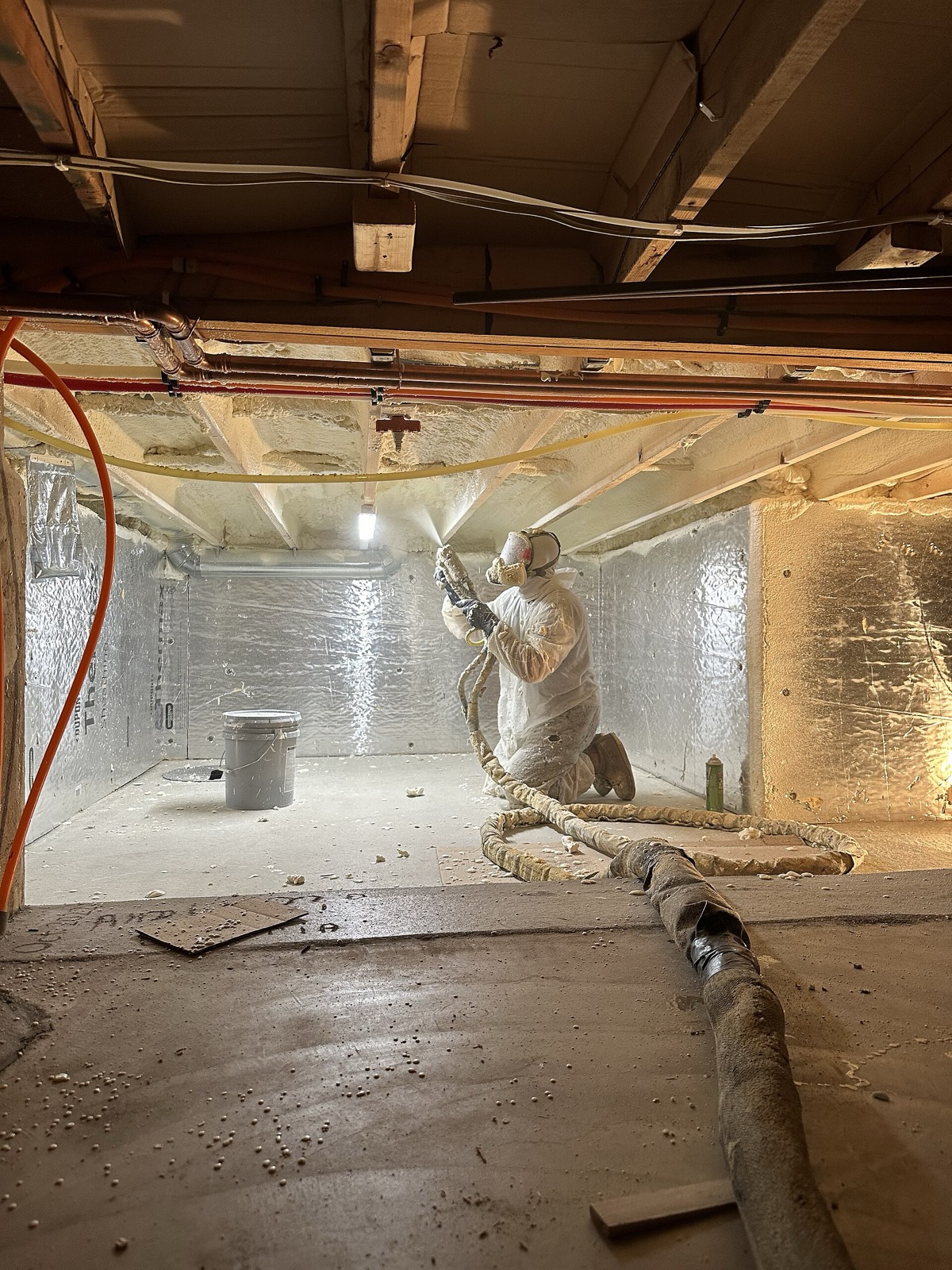 Worker in a protective suit spraying foam insulation in a crawl space lined with foil-faced foam board walls and a sealed floor