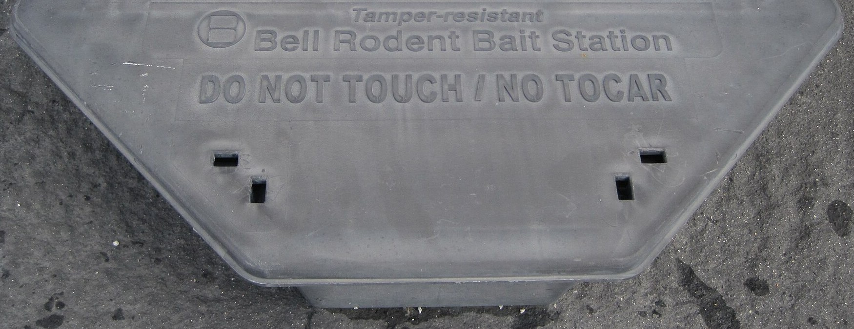 Gray plastic tamper-resistant rodent bait station on pavement, embossed with Do Not Touch