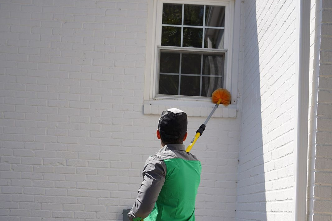 Vinx Pest Control technician using an extension duster to clear cobwebs from a window frame and exterior wall corner