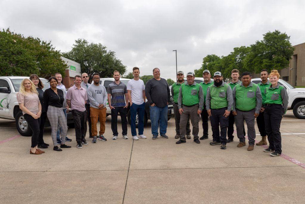 Group photo of the Vinx Pest Control team standing in a parking lot, with trucks in the background. Team members are dressed in green and gray uniforms, casual clothing, and business attire, showcasing a diverse and professional workforce.