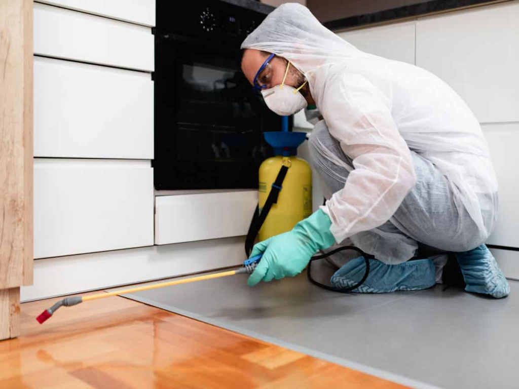 A pest control technician wearing protective gear, including a white suit, gloves, mask, and shoe covers, crouches in a kitchen while spraying pesticide along the baseboards with a long sprayer connected to a yellow canister.