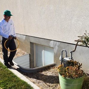 A pest control technician wearing a white shirt and green cap is standing outside a house holding a yellow hose, preparing to treat near a basement window well and gas meter.