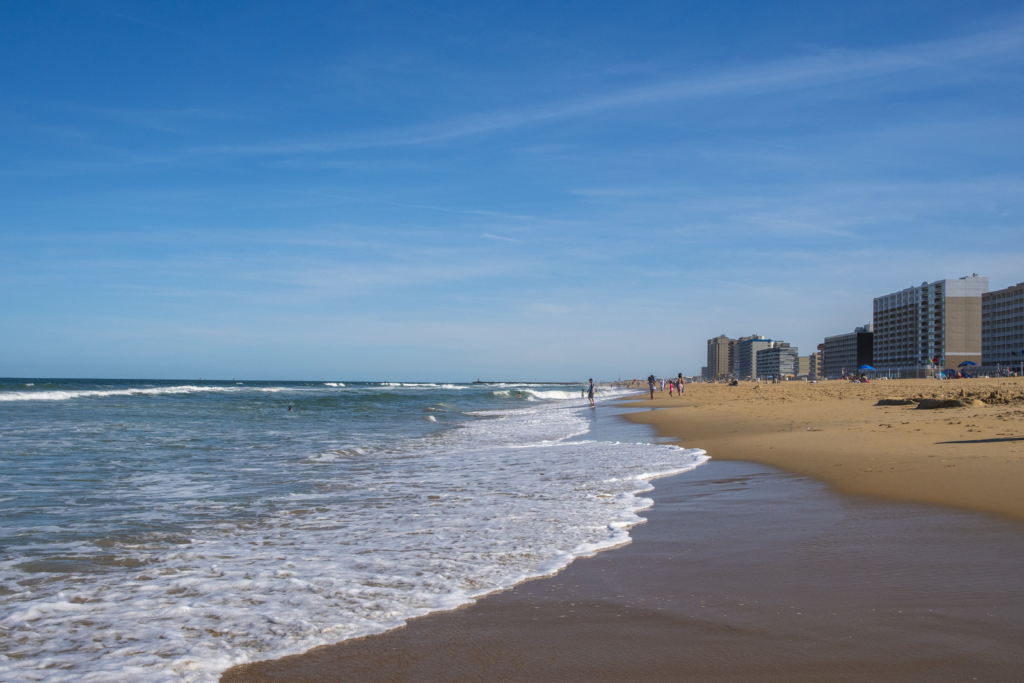 A wide view of Virginia Beach on a sunny day, showing gentle ocean waves washing onto the sandy shore. People are walking and relaxing along the beach, with tall beachfront buildings and hotels in the background under a clear blue sky.