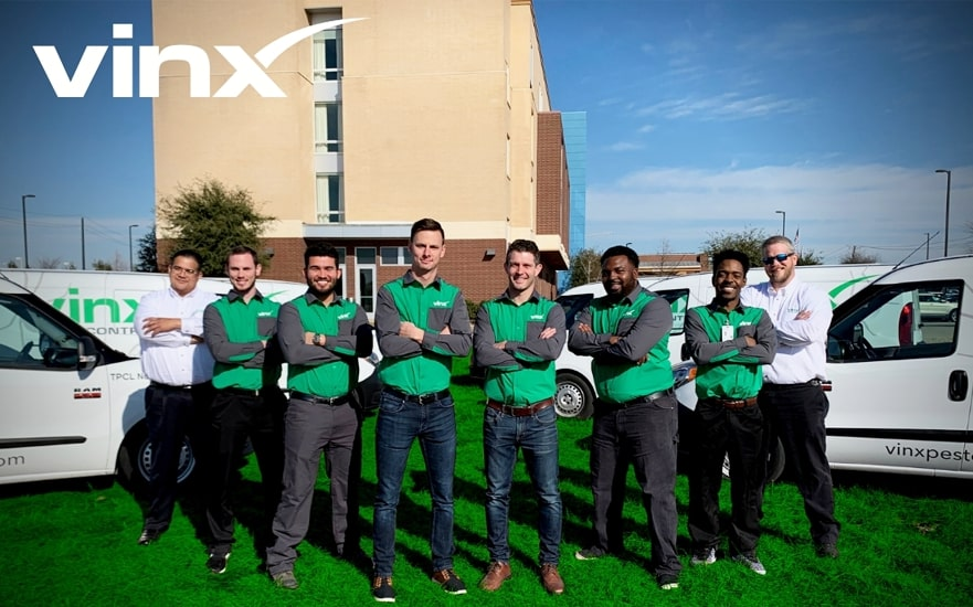 A group of eight pest control professionals from Vinx Pest Control stand confidently in front of their branded service vans and a building, all wearing matching green and gray uniforms. The Vinx logo is prominently displayed in the top left corner of the image.