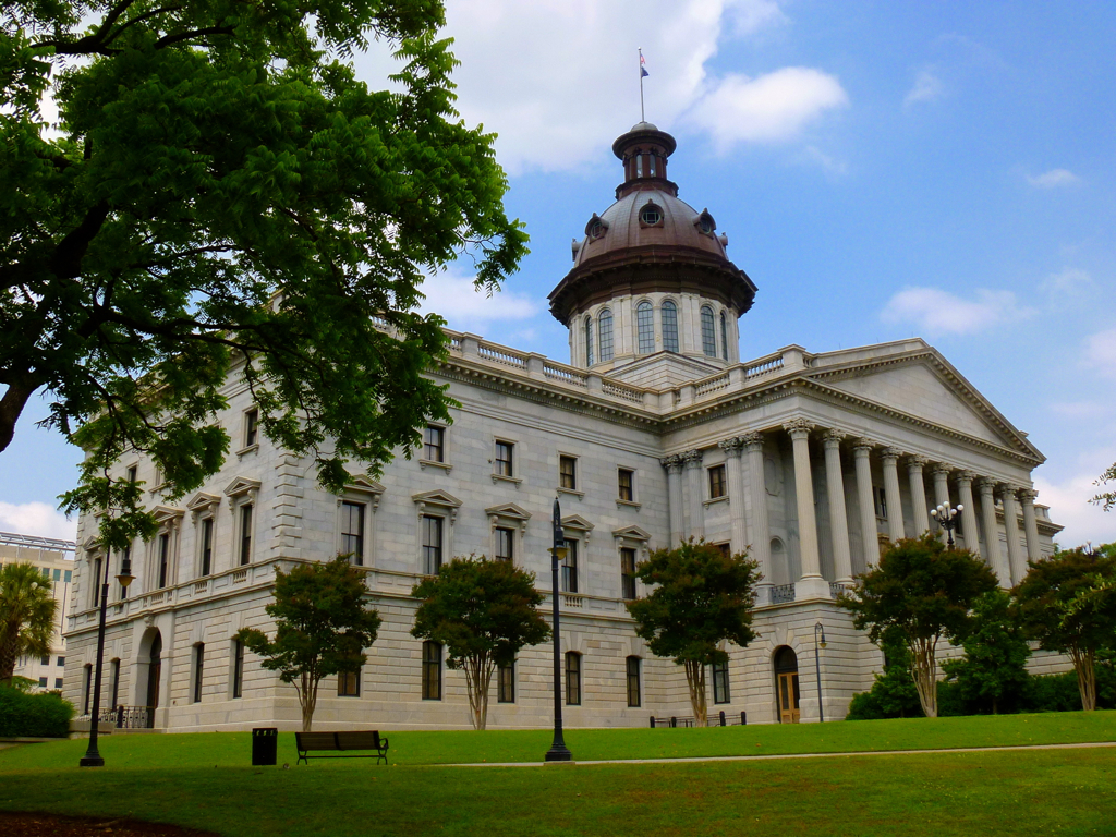A historic government building with neoclassical architecture, featuring tall white columns, arched windows, and a central copper dome topped with a flagpole. The structure is surrounded by a well-maintained lawn and trees under a partly cloudy sky.
