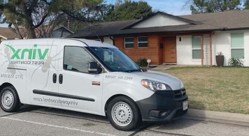 A white Vinx Pest Control service van parked on the street in front of a modern one-story home with a mix of white and wooden exterior siding. The van features the company's logo and website on the side.