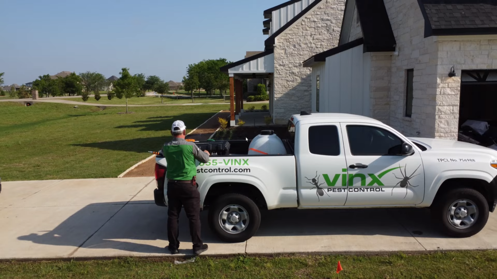 Vinx Pest Control technician standing behind a branded white pickup truck in a residential driveway, preparing pest control equipment outside a modern stone house on a sunny day. Have your home treated today in the Columbia and surrounding areas!